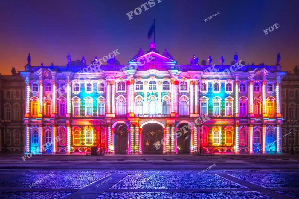 На фотографии изображён Эрмитаж, украшенный яркой и красочной подсветкой в рамках светового шоу. Роскошная архитектура здания подчёркнута игрой света и цветов, создавая праздничную атмосферу. Дворцовая площадь, выложенная брусчаткой, добавляет величия и уюта этому месту. Эрмитаж остаётся символом искусства и культуры Санкт-Петербурга, привлекая внимание жителей и туристов.